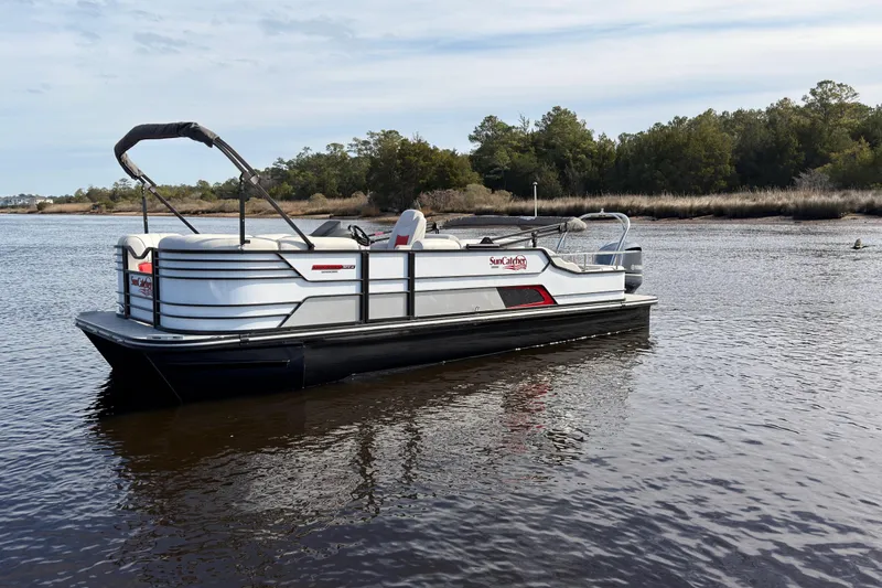 Slide: The Image of 2025 SunCatcher ELITE SERIES 324 SS pontoon boat on calm water, surrounded by trees. - 2