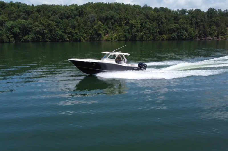 Slide: The Image of 2021 Scout 330 LXF boat cruising on a serene lake with lush green forest backdrop. - 18