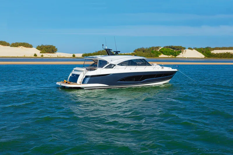 Slide: The Image of 2026 Riviera 5600 Sport Yacht Platinum Edition cruising on blue waters near sandy shore. - 6