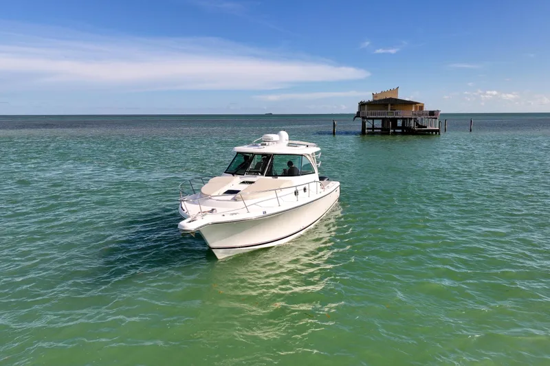 Slide: The Image of 2016 Pursuit OS 385 Offshore boat cruising near a stilt house on calm, turquoise waters. - 11