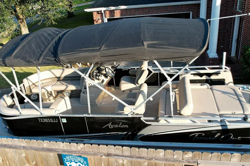 Slide: The Image of 2021 Avalon LSZ 2485 pontoon boat with black canopy and beige seating, parked near a house. - 2