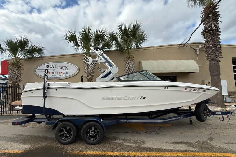 The Image of 2022 MasterCraft XT24 boat on trailer at dealership with palm trees. - 0