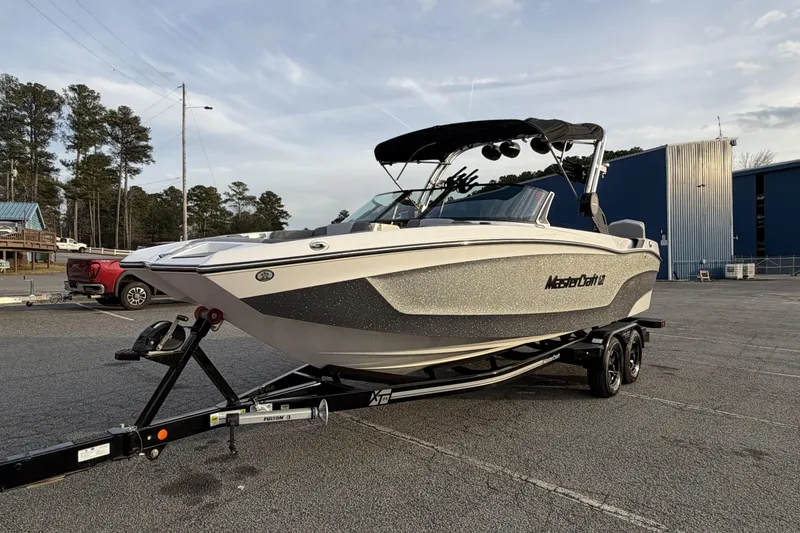 Slide: The Image of 2023 MasterCraft XT24 boat on trailer in parking lot, surrounded by trees and buildings. - 7