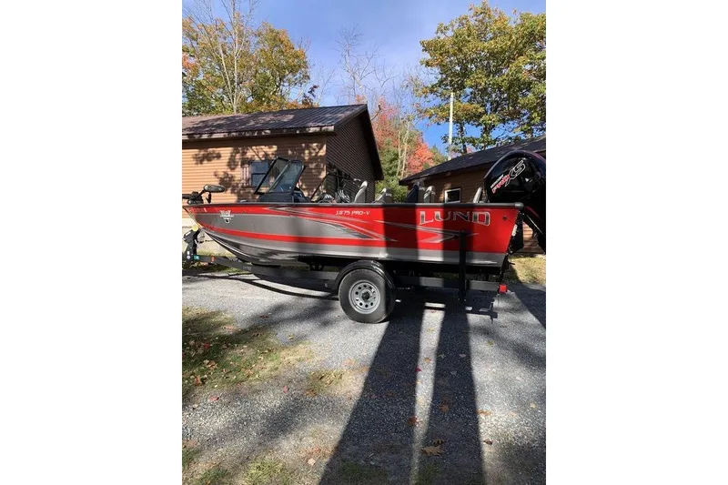 The Image of 2022 Lund 1875 Pro-V Sport boat on trailer, parked near a house with autumn trees. - 0