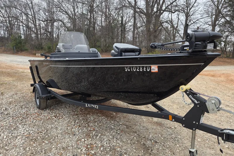 Slide: The Image of 2023 Lund 1650 Rebel XL Sport boat on trailer, parked on gravel, surrounded by trees. - 2