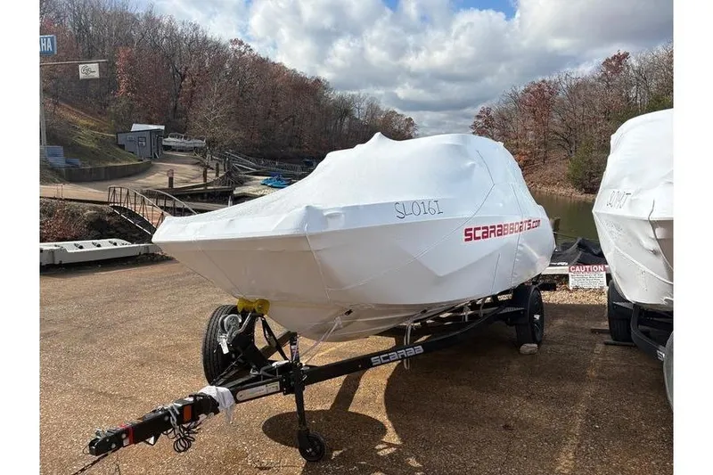 Slide: The Image of 2026 Scarab 210 SE boat covered on trailer, parked outdoors near a lake. - 10