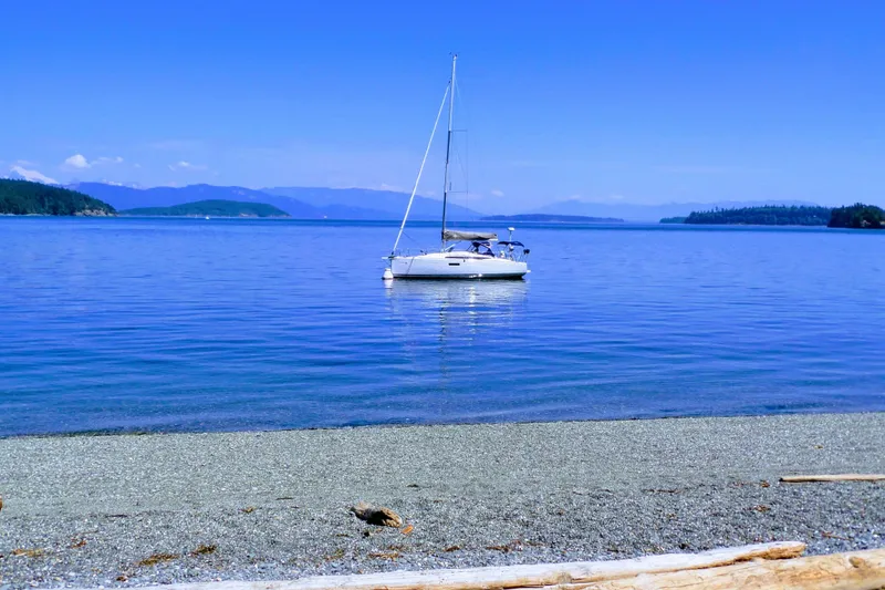 Slide: The Image of 2017 Jeanneau Sun Odyssey 349 sailboat anchored in serene blue waters near a pebble beach. - 23