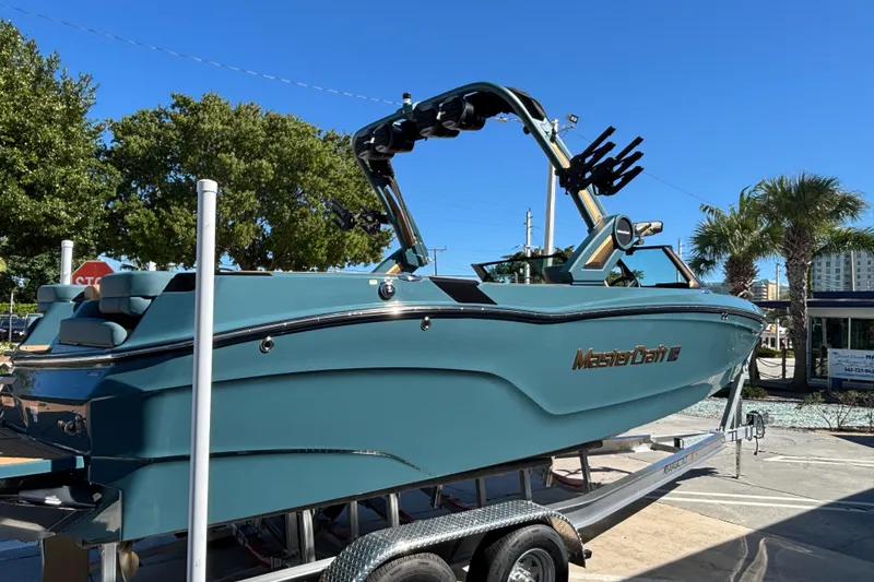 Slide: The Image of 2026 MasterCraft XT24 boat on trailer, teal color, sunny day, palm trees in background. - 2