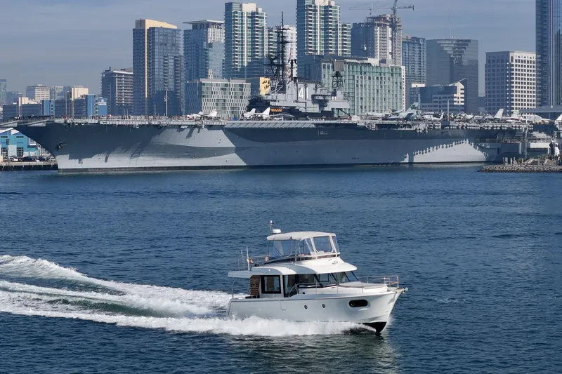 Slide: The Image of 2025 Beneteau Swift Trawler 41 cruising near a large battleship in an urban harbor. - 74