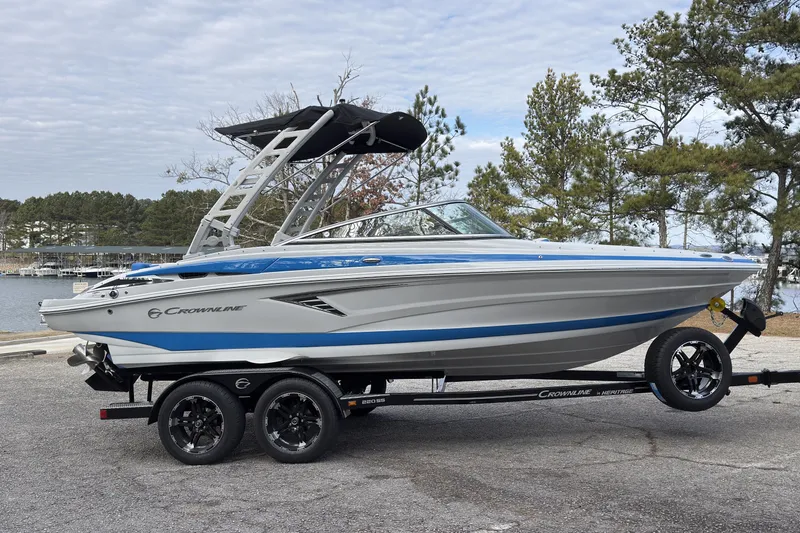 Slide: The Image of 2026 Crownline 220 SS boat on trailer, parked near a lake with trees in the background. - 2