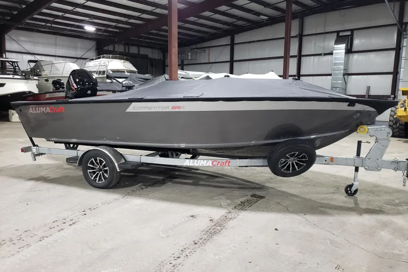 The Image of 2026 Alumacraft Competitor 185X Tiller boat on trailer in indoor storage. - 0