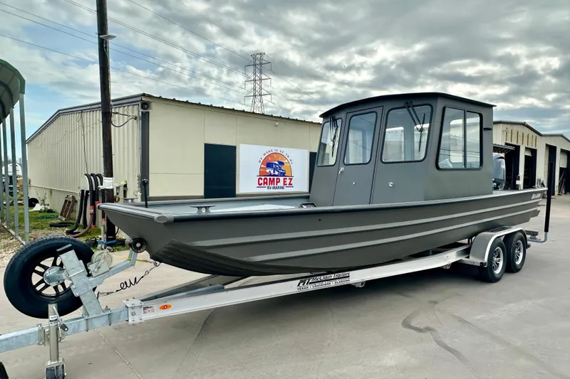 The Image of 2026 SeaArk 2472 CUB boat on trailer, parked outside a warehouse under cloudy skies. - 1