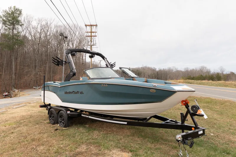 The Image of 2026 MasterCraft NXT23 boat on trailer, parked roadside with forest background. - 0