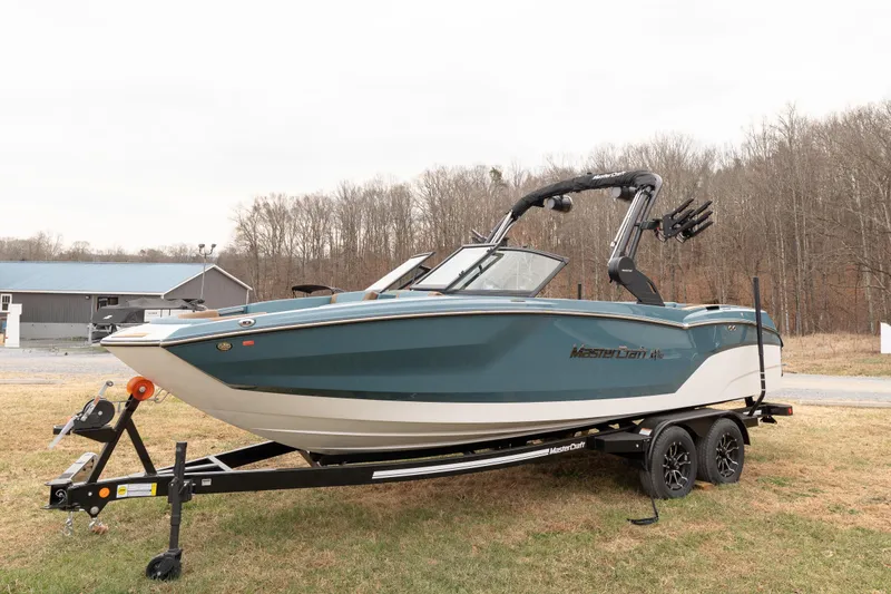 Slide: The Image of 2026 MasterCraft NXT23 boat on trailer, parked on grass with wooded background. - 2