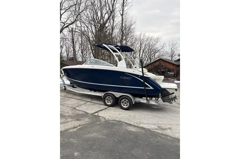 Slide: The Image of 2015 Cobalt R5 boat on trailer, parked outdoors, with trees and a wooden building in the background. - 2