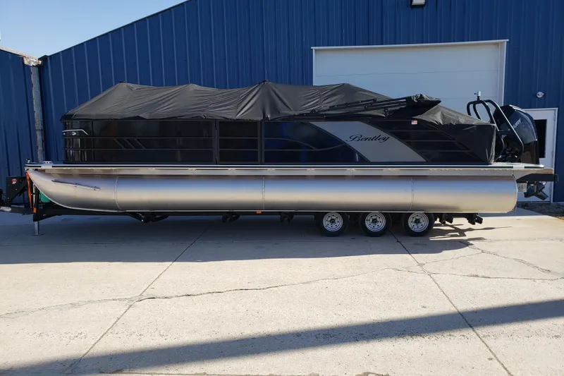 Slide: The Image of 2026 Bentley Pontoons Elite 253 Swingback Bench, covered, parked outside a blue building. - 5