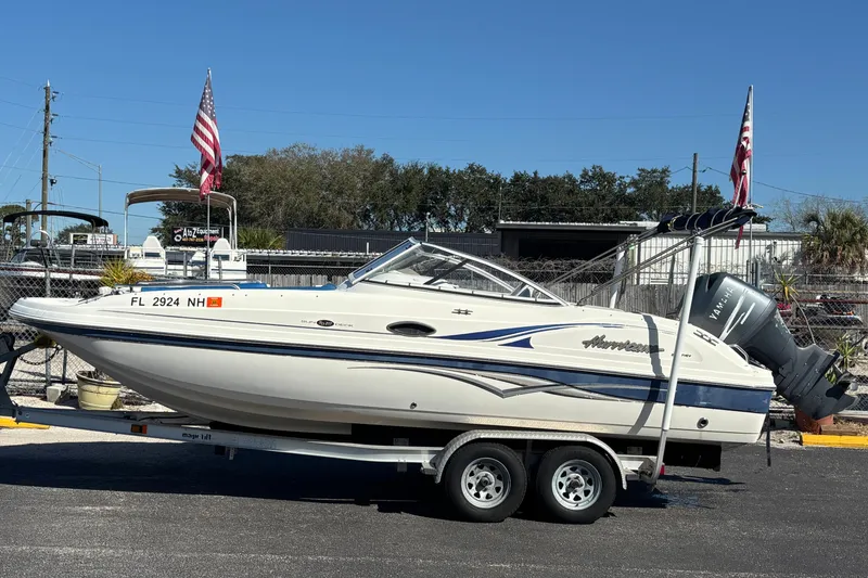 The Image of 2006 Hurricane SunDeck 217 OB boat on trailer with flags, parked outdoors. - 0