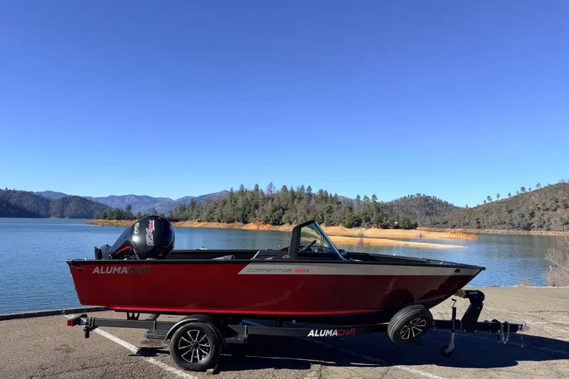 The Image of 2025 Alumacraft Competitor 185X Sport boat on trailer by scenic lake and mountains. - 5