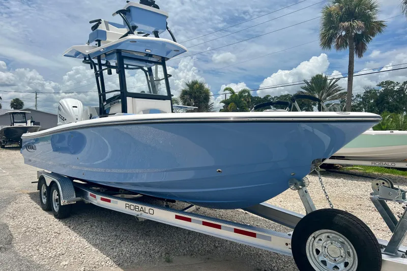 The Image of 2025 Robalo 266 Cayman SD boat on trailer, blue hull, palm trees in background. - 0