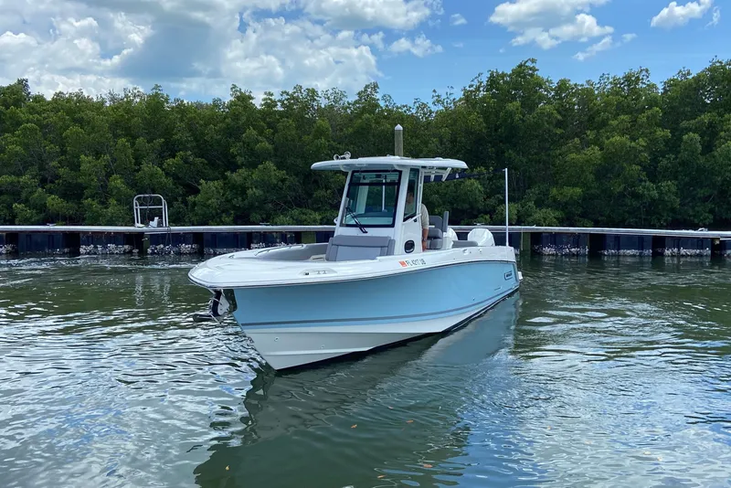 Slide: The Image of 2025 Boston Whaler 280 Outrage boat on calm water near a dock, surrounded by lush greenery. - 6