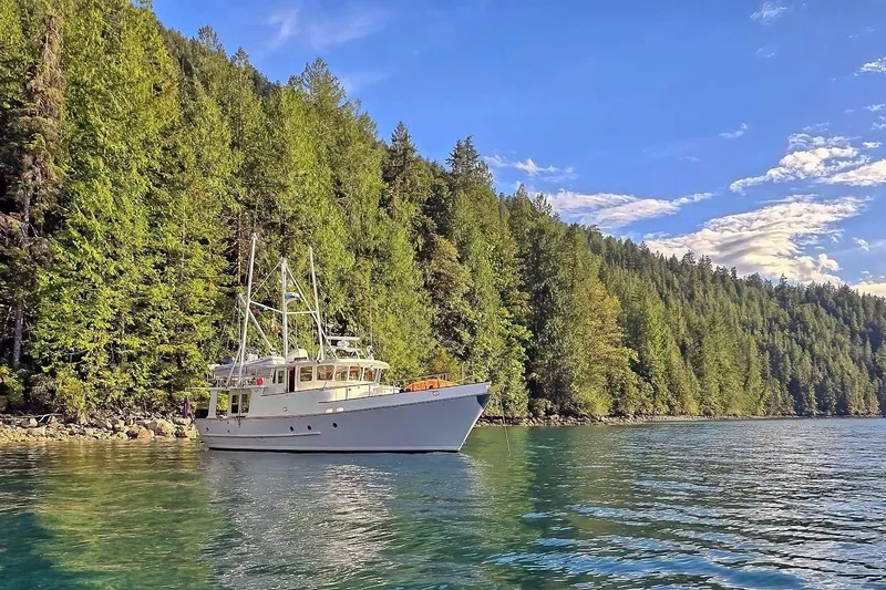 Slide: The Image of Nordhavn 46 yacht from 2001 cruising near lush forested shoreline under clear blue sky. - 70