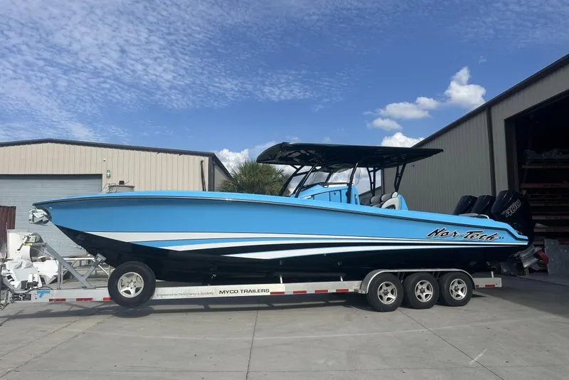 Slide: The Image of 2025 Nor-Tech 340 Sport Center Console boat on trailer, vibrant blue, parked outside warehouse. - 2