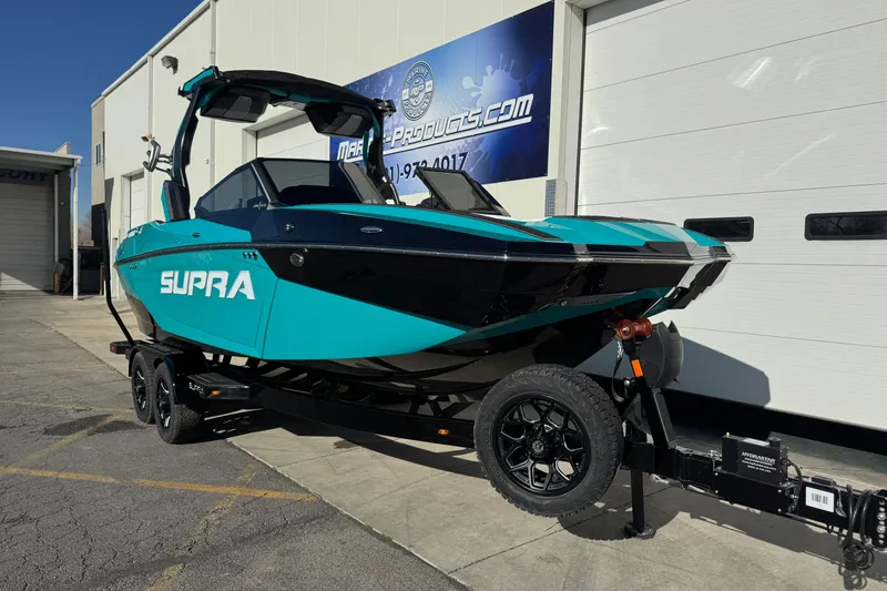 Slide: The Image of 2026 Supra SA 575 boat in teal and black, parked on a trailer outside a building. - 2