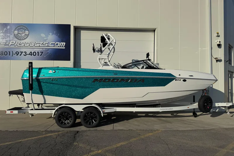Slide: The Image of 2026 Moomba Tykon boat on trailer, teal and white design, parked outside a building. - 2