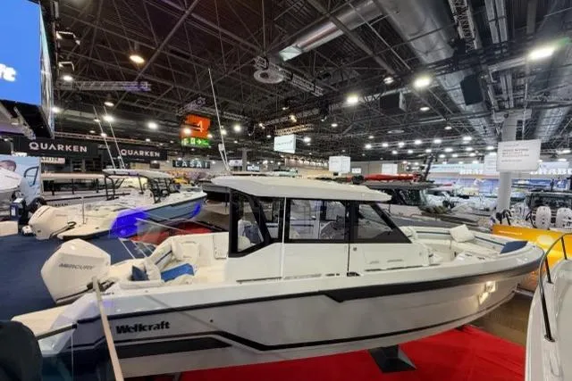 The Image of 2026 Wellcraft 28 Explorer boat displayed at indoor exhibition, surrounded by other boats. - 0