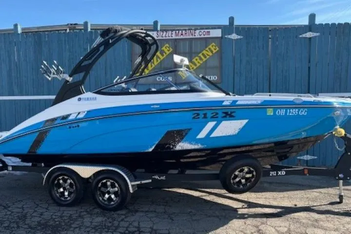 The Image of 2021 Yamaha Boats 212XD in blue, parked on a trailer at a marina. - 0