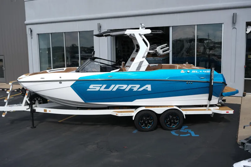 Slide: The Image of 2026 Supra SL boat in blue and white on a trailer outside a building. - 2