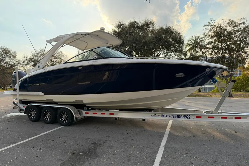 The Image of 2019 Regal 29 OBX boat on trailer in parking lot, surrounded by trees. - 1