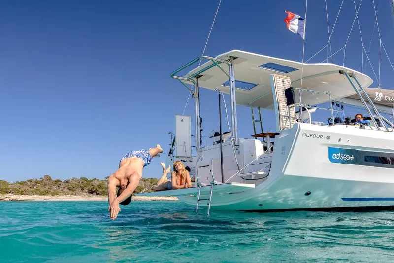 Slide: The Image of Man diving off 2026 Dufour 48 yacht, woman relaxing, clear blue water. - 26