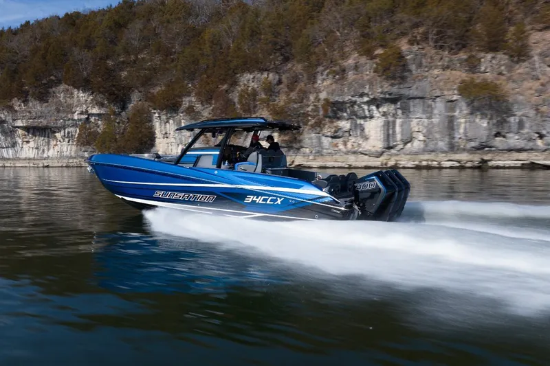 Slide: The Image of 2026 Sunsation 34 CCX speedboat cruising on a lake with rocky shoreline backdrop. - 34