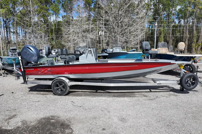Slide: The Image of 2026 Xpress H22B Bay boat in red, parked on a trailer, surrounded by trees. - 1