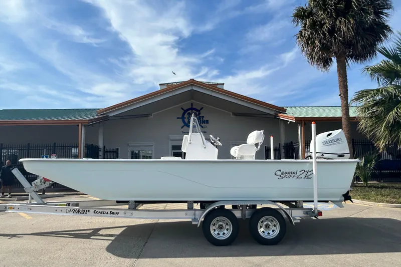 The Image of 2026 Coastal Skiff 212 boat on trailer, parked outside a building with palm trees. - 1