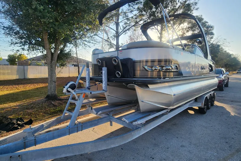 Slide: The Image of 2023 Avalon Excalibur 25' pontoon boat on trailer, parked outdoors in sunny setting. - 2