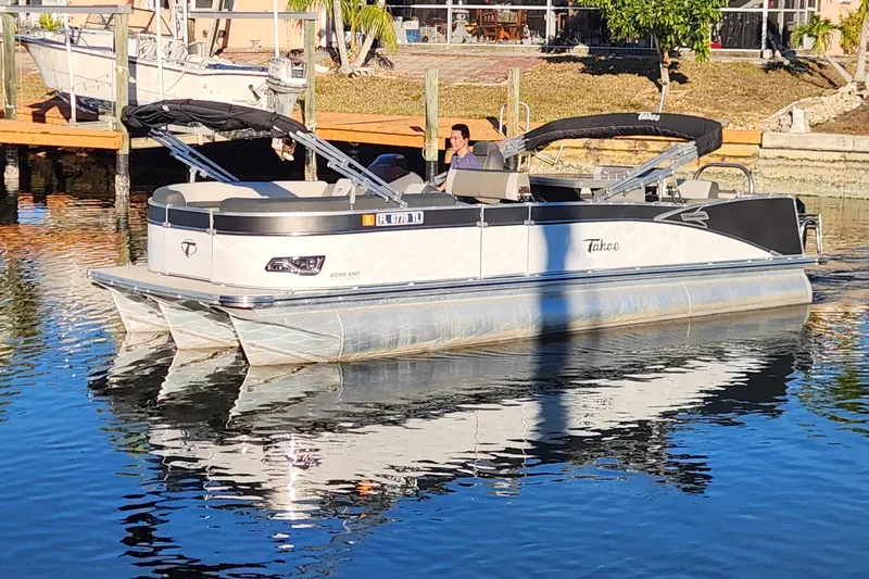 Slide: The Image of 2022 Tahoe Tritune 2585 ENT pontoon boat on calm water near a dock. - 2