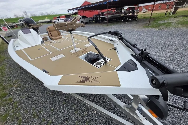 Slide: The Image of 2026 Xpress H20CC boat with tan deck, parked on gravel near red buildings. - 22