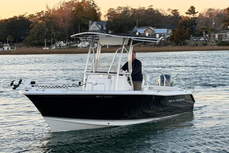Slide: The Image of 2014 Sea Hunt Ultra 211 boat on water, with a person steering, near a shoreline. - 12