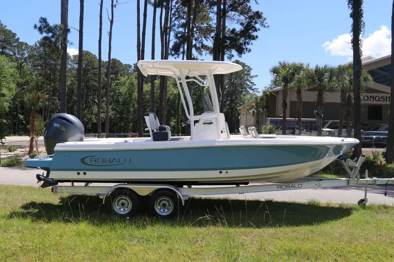 The Image of 2026 Robalo 226 Cayman boat on trailer, parked outdoors near trees and building. - 0