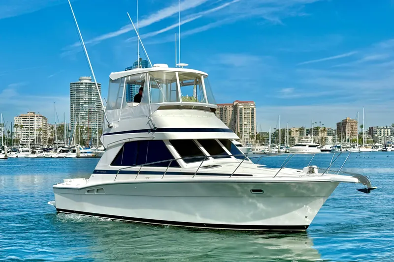 Slide: The Image of 1998 Riviera 34 Convertible yacht cruising in a marina with city skyline backdrop. - 2