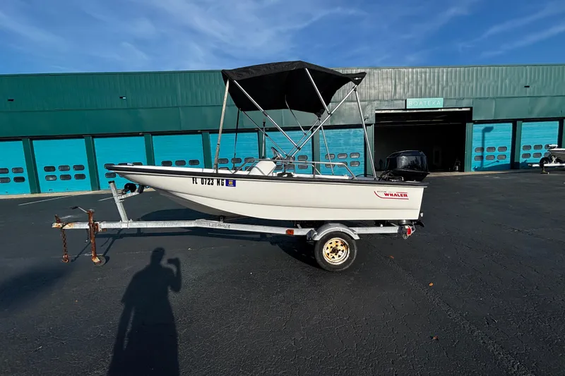 The Image of 2000 Boston Whaler Sport 13 boat on trailer, parked outside a teal building. - 1