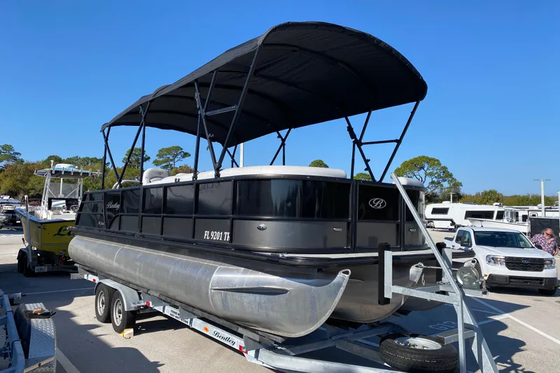 Slide: The Image of 2024 Bentley Pontoons Legacy Swingback on trailer, black canopy, parked outdoors. - 2