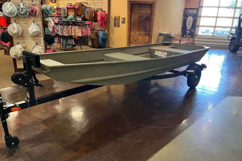 The Image of 2026 Tracker Grizzly 14 Jon boat on trailer in showroom, surrounded by clothing displays. - 0