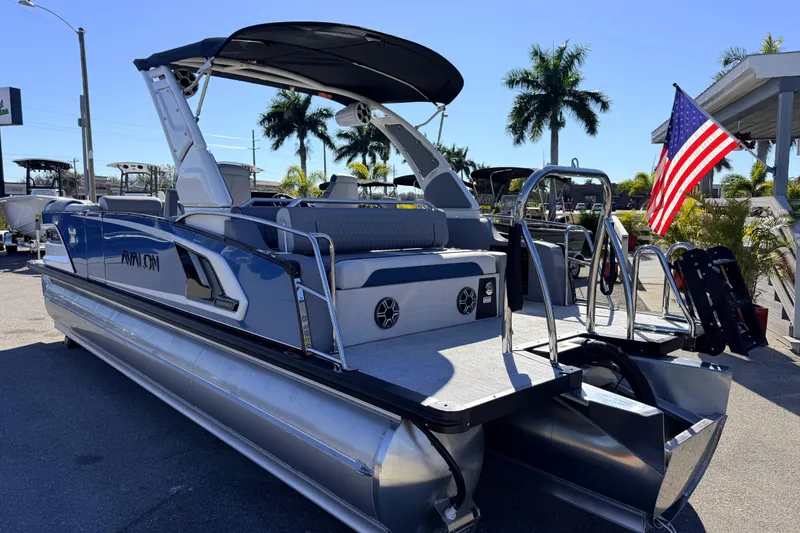 Slide: The Image of 2026 Avalon 2585 Excalibur LTD Flex Lounge Tri Toon boat with American flag, parked outdoors. - 9