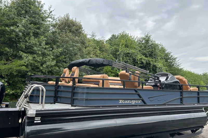 Slide: The Image of 2026 Ranger 223F pontoon boat with tan seating, parked near lush green trees. - 1