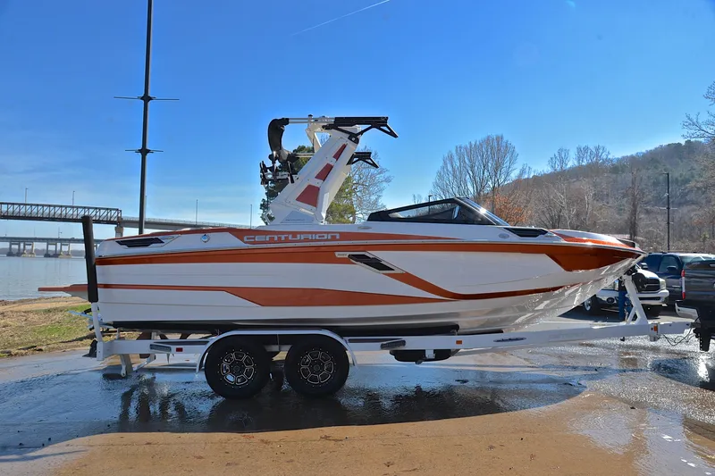 Slide: The Image of 2023 Centurion Ri230 boat on trailer, orange and white design, parked near a river. - 17