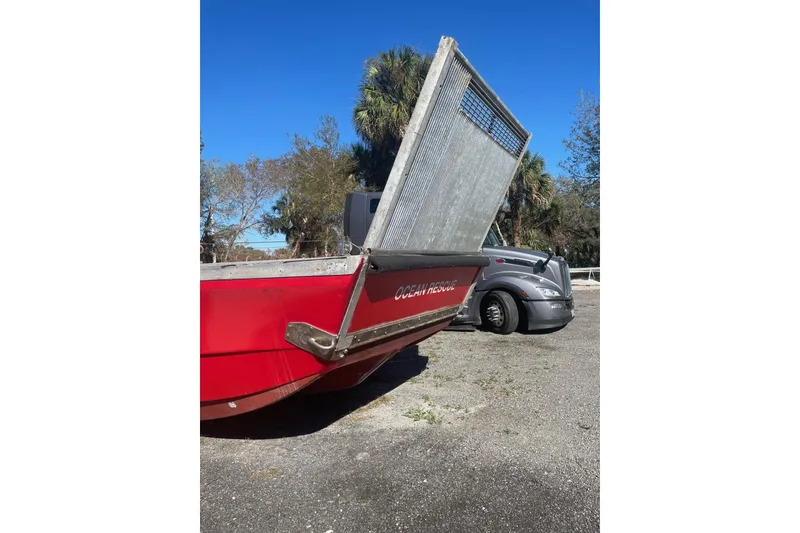 Slide: The Image of Red 1987 Commercial Buoy Tender with open ramp, parked near a truck, surrounded by trees. - 24
