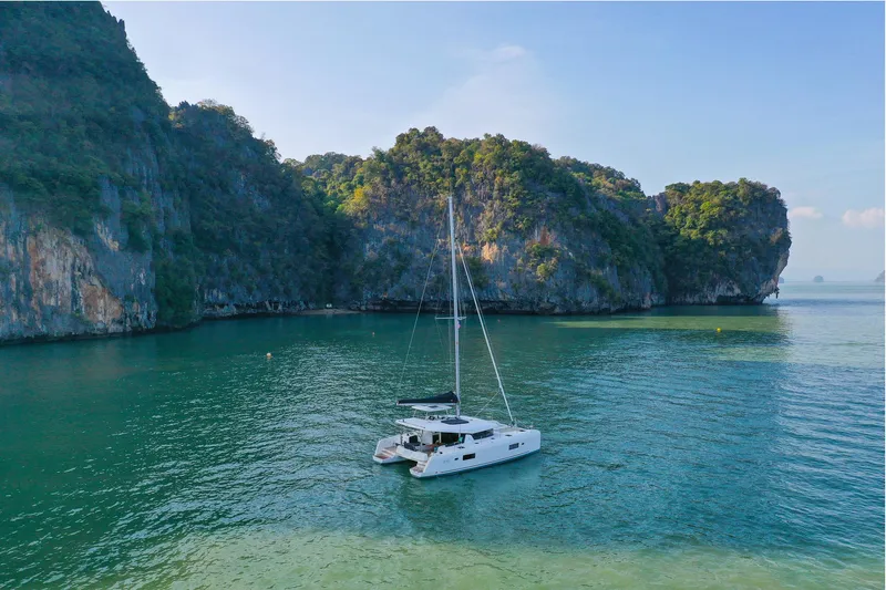 Slide: The Image of A 2025 Lagoon 42 Millenium catamaran sailing in a serene, tropical bay with lush cliffs. - 0